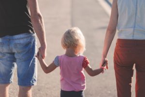 Young child walking between 2 adults, one male and one female. All are holding hands. Picture taken from behind.