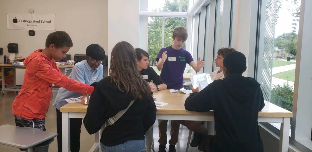 Youth gesturing and discussing robotics as they sit or stand at a table