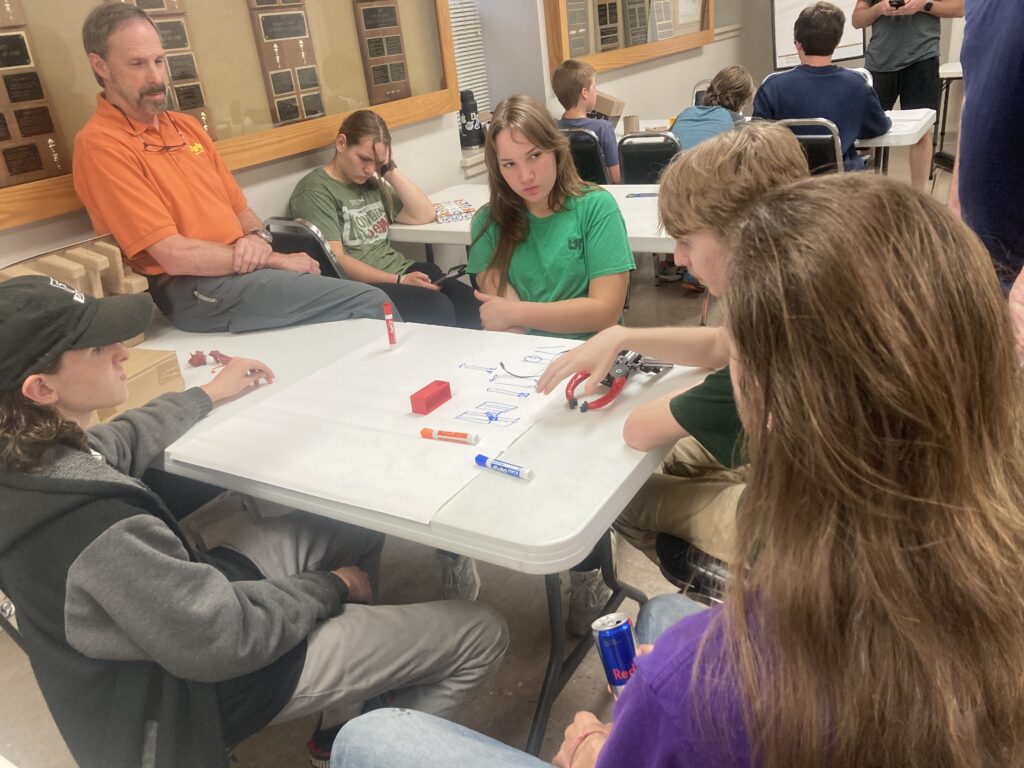 A group of robot team members sit around a table while discussing robotics and sketching robot designs on a piece of paper